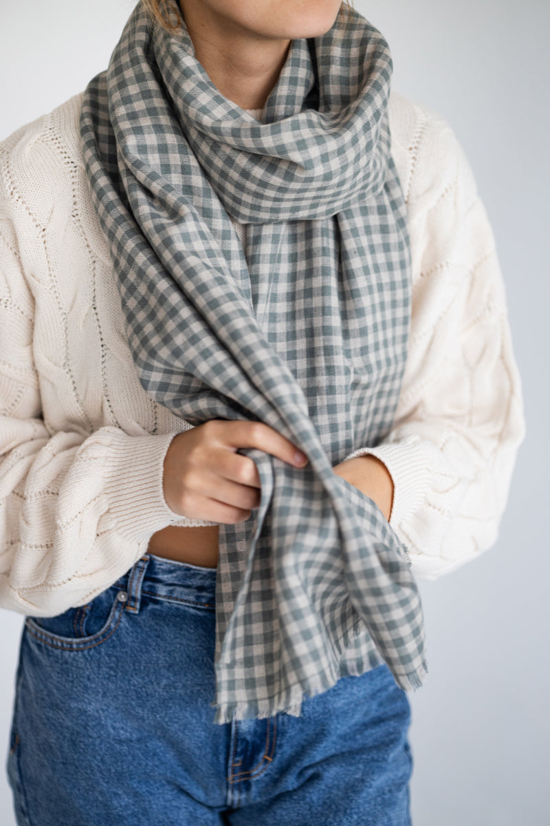 Person wearing a plaid linen scarf, white sweater, and blue jeans on a light gray background
