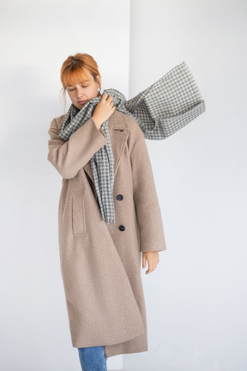 Woman wearing a beige coat with a plaid linen scarf against a white background

