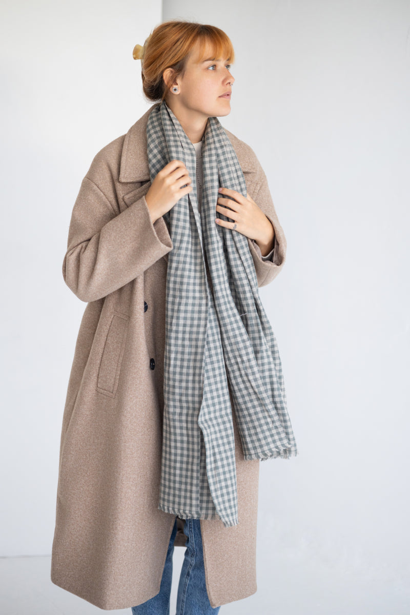Woman wearing a beige coat with a plaid linen scarf against a white background
